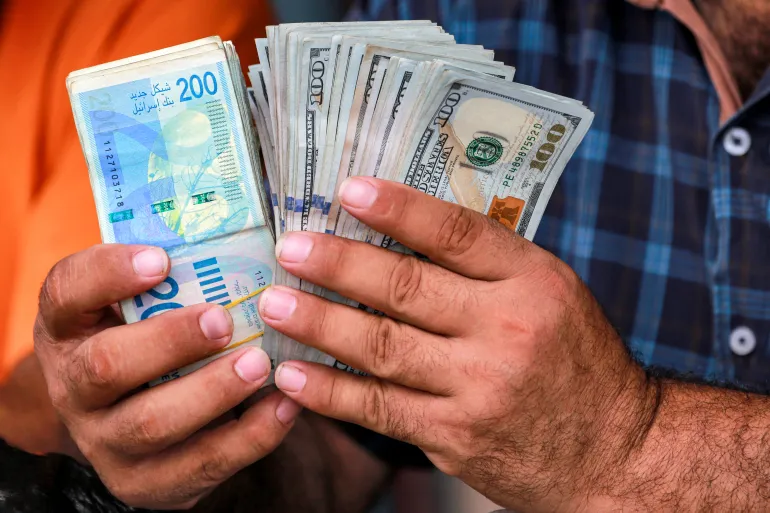 A man counts stacks of Israeli shekel and US dollar banknotes at an informal money exchange stall in Deir el-Balah in the central Gaza Strip on July 2, 2024 amid the ongoing conflict in the Palestinian territory between Israel and Hamas. (Photo by Bashar TALEB / AFP)
المصارف الفلسطينية لا تستطيع الوصول مباشرة إلى نظام المدفوعات الإسرائيلي (الفرنسية)